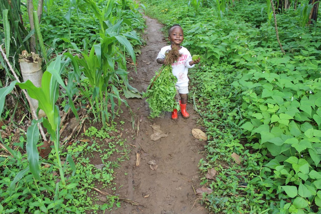 Young Prince on a farm path in Masaka holding plants and fruit